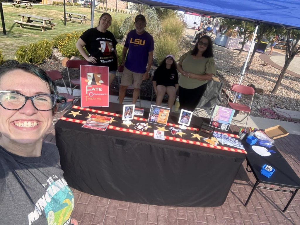 A Fate Community Theatre member smiles in front of the company's event table while volunteers talk with visitors