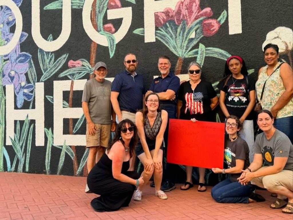 Fate Community Theatre members gather in front of the Fate mural, holding a handmade Fate Community Theater sign